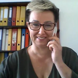 Uśmiechnięta kobieta w okularach rozmawia przez telefon na tle regału z segregatorami. Widok z bliska, jasne oświetlenie.