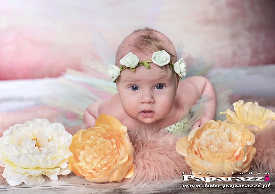 Niemowlę w tiulowej spódniczce i wianku z białych róż, leżące na puszystym różowym dywaniku, otoczone żółtymi piwoniami na tle rozmytego różowo-białego tła.