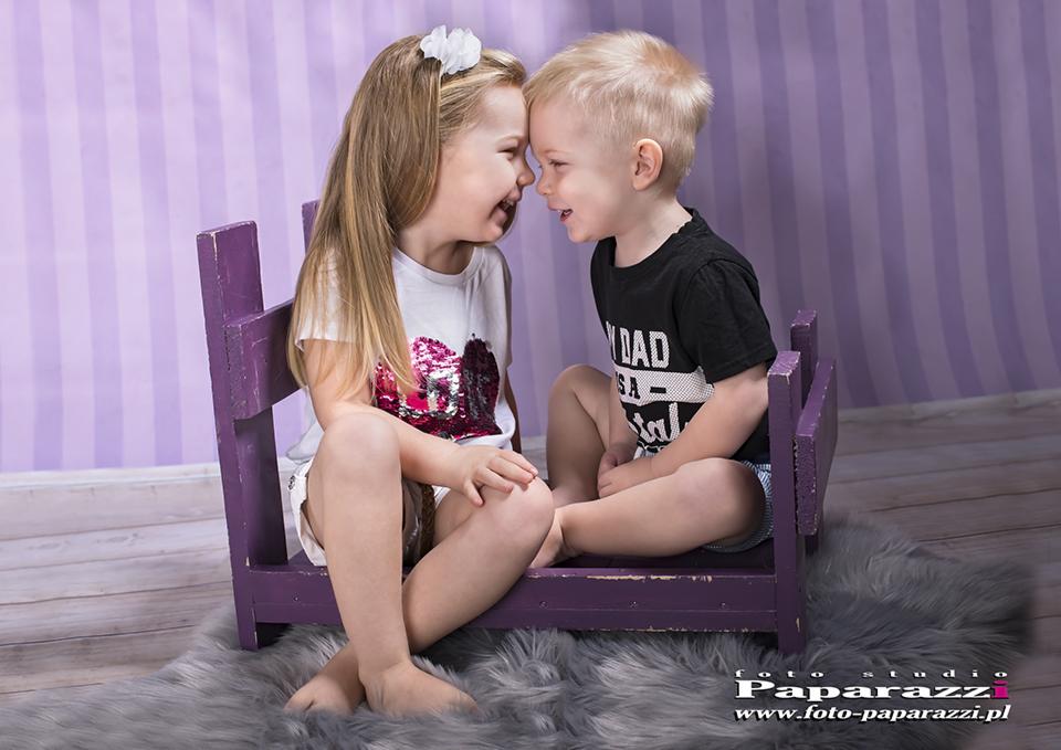 Uśmiechnięta dziewczynka z kwiatem we włosach i chłopiec siedzą na małym fioletowym krześle, stykając się czołami na tle fioletowej ściany w paski.