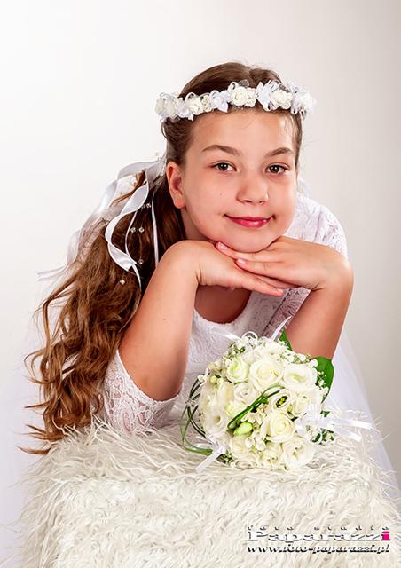 Dziewczynka w białej sukience z wiankiem na głowie, opiera brodę na dłoniach, trzymając bukiet białych róż.