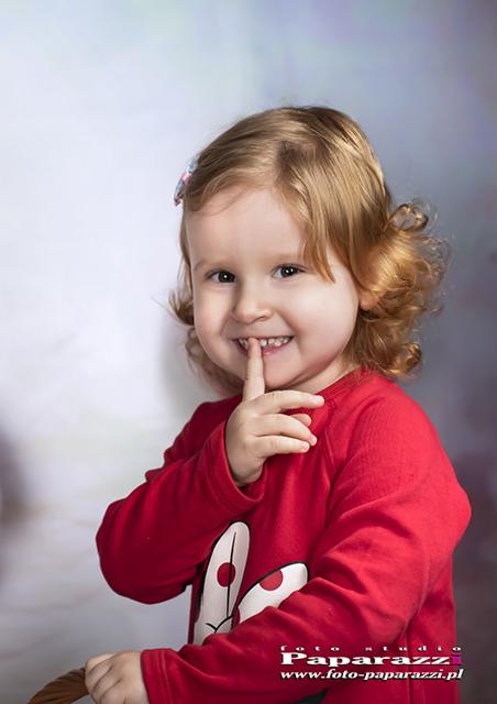 Uśmiechnięta dziewczynka z kręconymi blond włosami, w czerwonej bluzce z motywem uszu królika, trzyma palec na ustach na rozmytym tle.