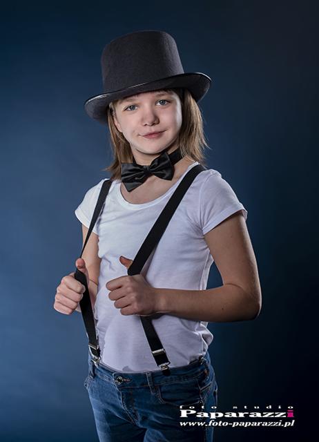 Portret młodej osoby w cylindrze, muszce i szelkach na tle ciemnego studia, stylizacja nawiązująca do klasyki.