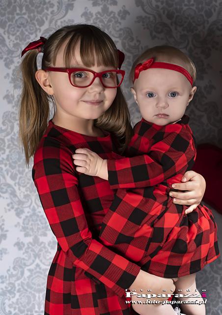Dwie dziewczynki, starsza trzyma młodszą na rękach, obie ubrane w czerwono-czarne sukienki w kratę, na głowach mają czerwone kokardki, starsza ma czerwone okulary, tło stanowi wzorzysta tapeta.