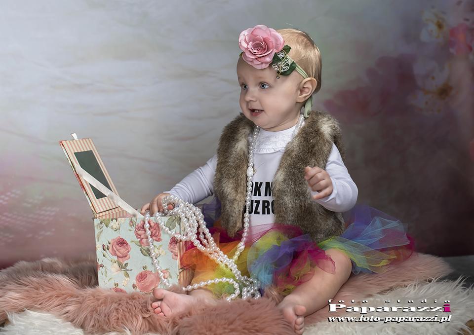 Dziewczynka w futrzanej kamizelce i tiulowej spódniczce siedzi na futrze, bawi się perłami wyciągniętymi z pudełka w kwiaty, na głowie ma opaskę z różą.