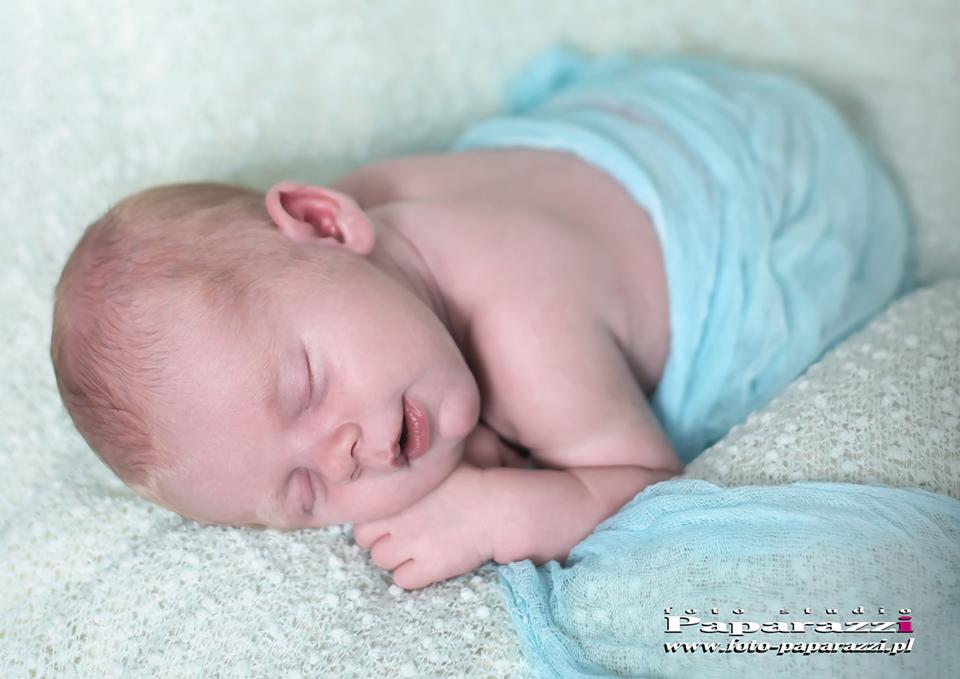 Śpiący noworodek owinięty w błękitny materiał, ułożony na miękkim, teksturowanym tle.