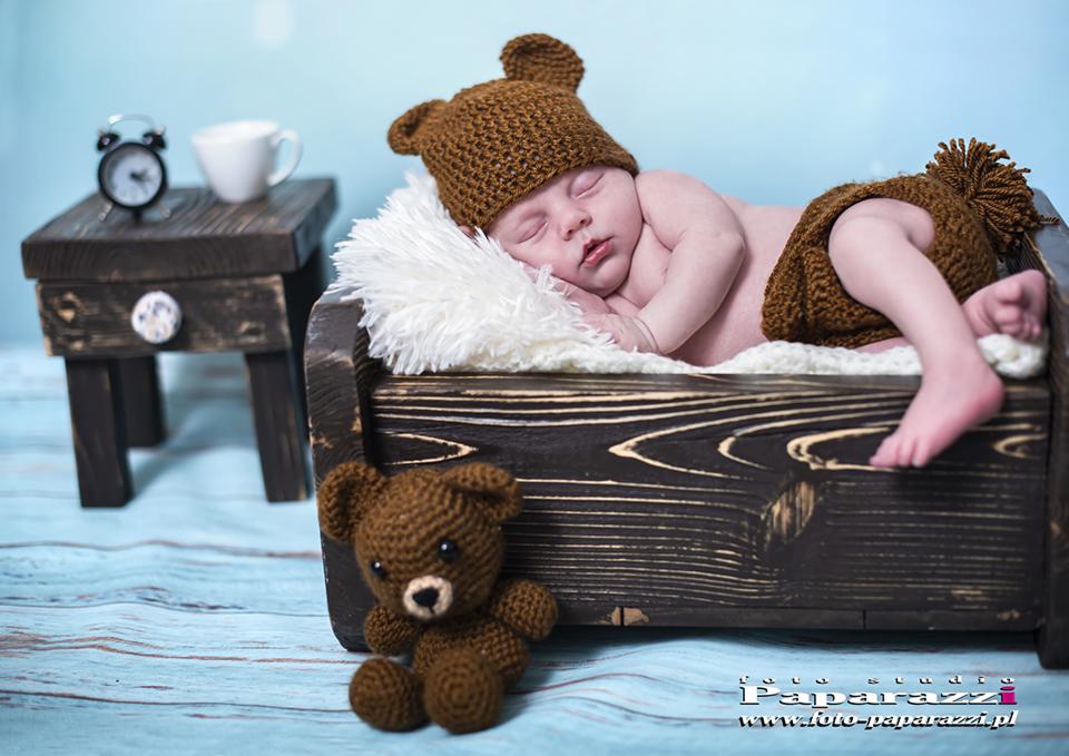 Śpiący noworodek w brązowym stroju misia, leżący w małym drewnianym łóżeczku na białym futerku, obok siedzi pluszowy miś, a w tle stoi mały stolik z budzikiem i kubkiem.