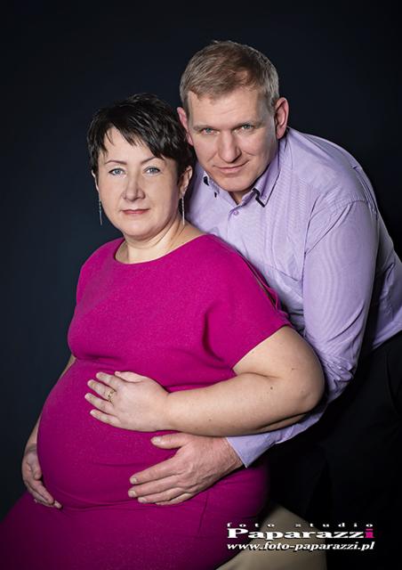 Portret kobiety w różowej sukience ciążowej, z dłońmi na brzuchu, obok stoi mężczyzna w liliowej koszuli, oboje patrzą w obiektyw na ciemnym tle.