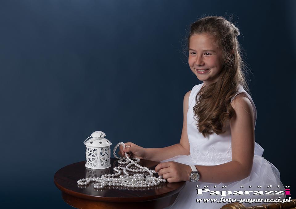 Uśmiechnięta dziewczynka w białej sukience, trzymająca sznur pereł, obok biały lampion na okrągłym stoliku, na ciemnoniebieskim tle.