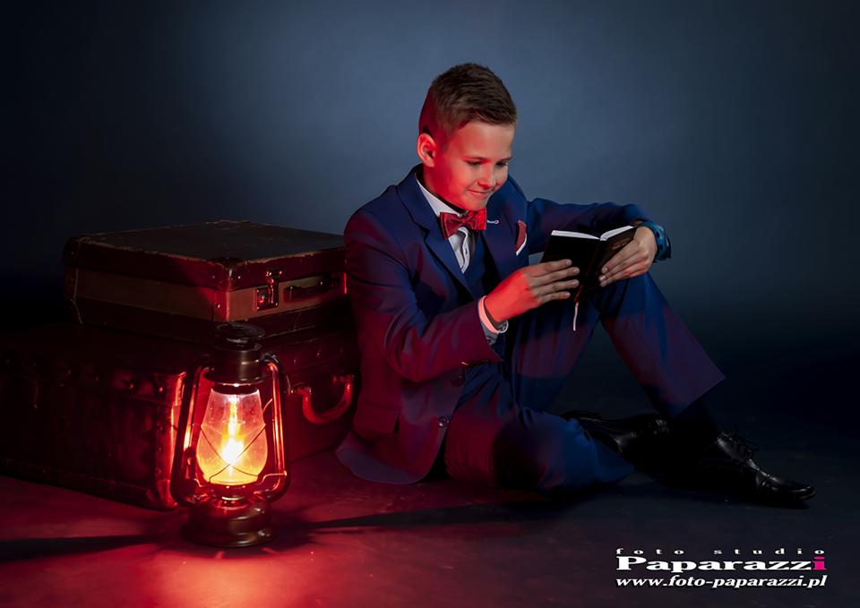 Chłopiec w granatowym garniturze z muchą siedzi na podłodze, czytając książkę przy naftowej lampie i starych walizkach w ciemnym studiu.