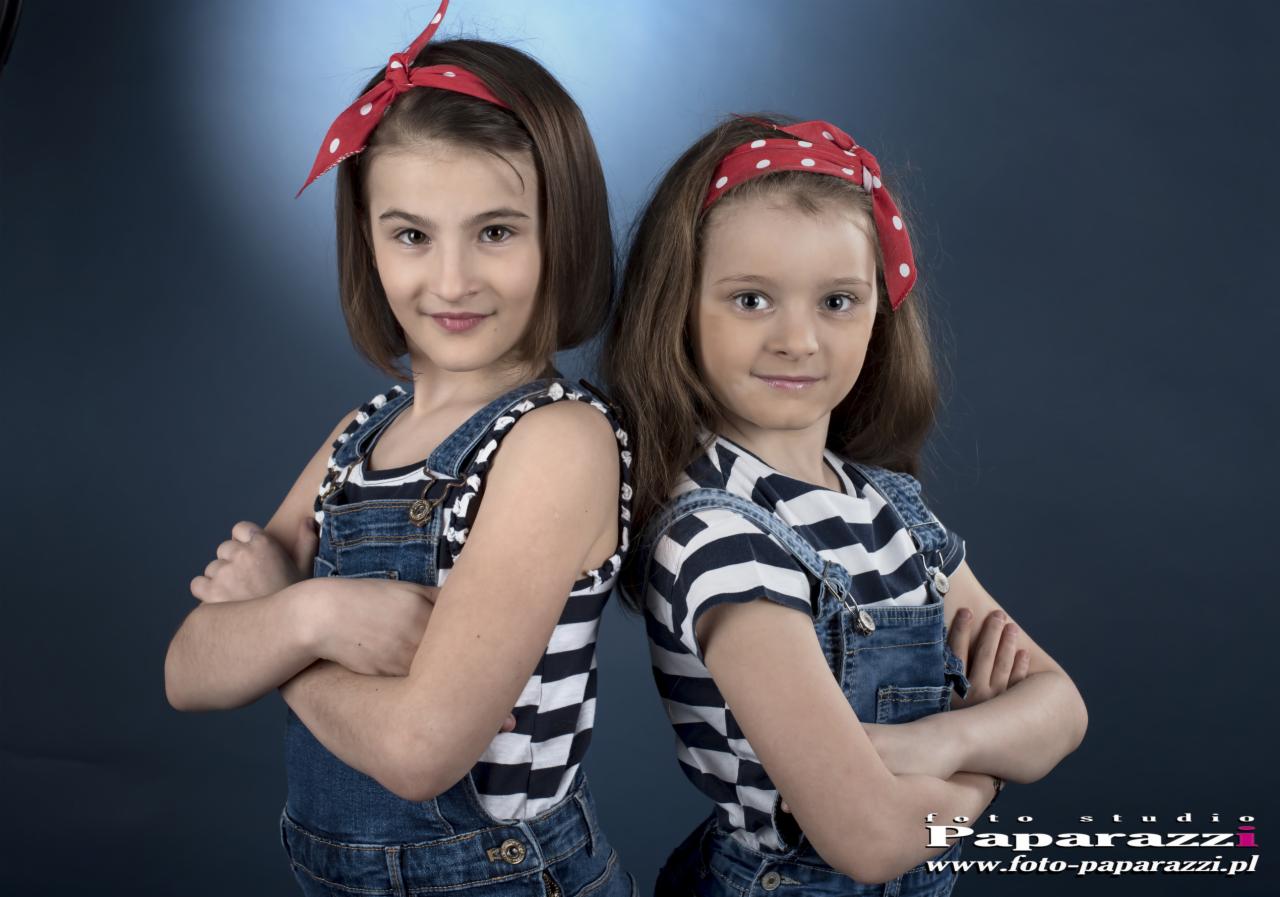 Portret studyjny dwóch uśmiechniętych dziewczynek w dżinsowych ogrodniczkach i bluzkach w paski, z czerwonymi opaskami we włosach, stojących plecami do siebie na tle granatowego tła.