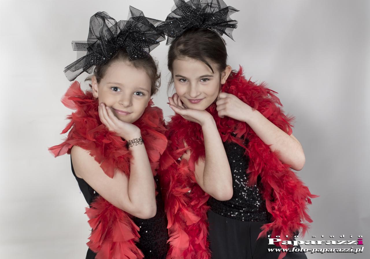 Portret dwóch uśmiechniętych dziewczynek w czarnych sukienkach z cekinami, czerwonych boa z piór i czarnych ozdobach do włosów z tiulu i brokatu, pozujących w studio.