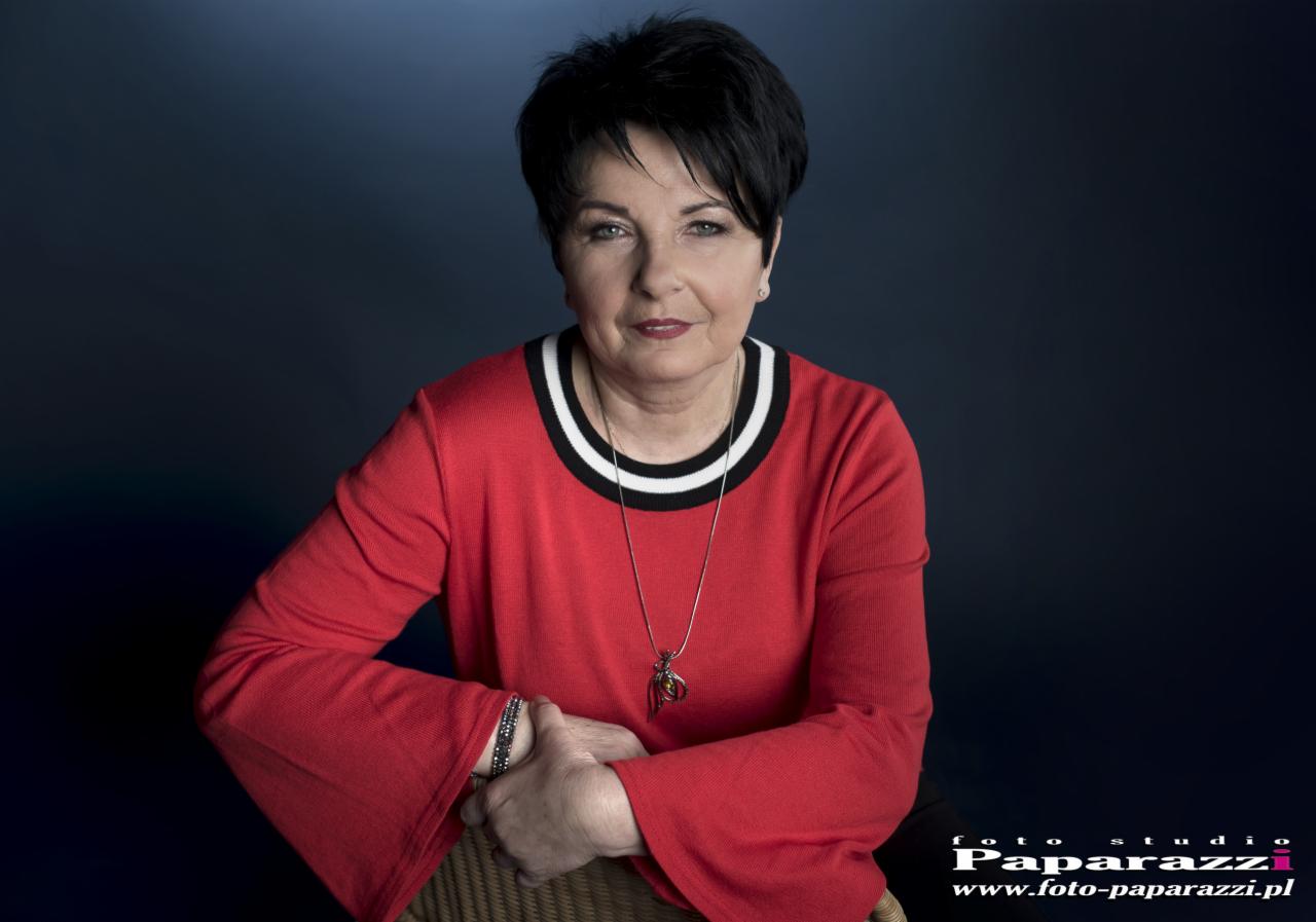 Portret kobiety w czerwonej bluzce z czarno-białym dekoltem, na ciemnym tle, z delikatnym uśmiechem i biżuterią.