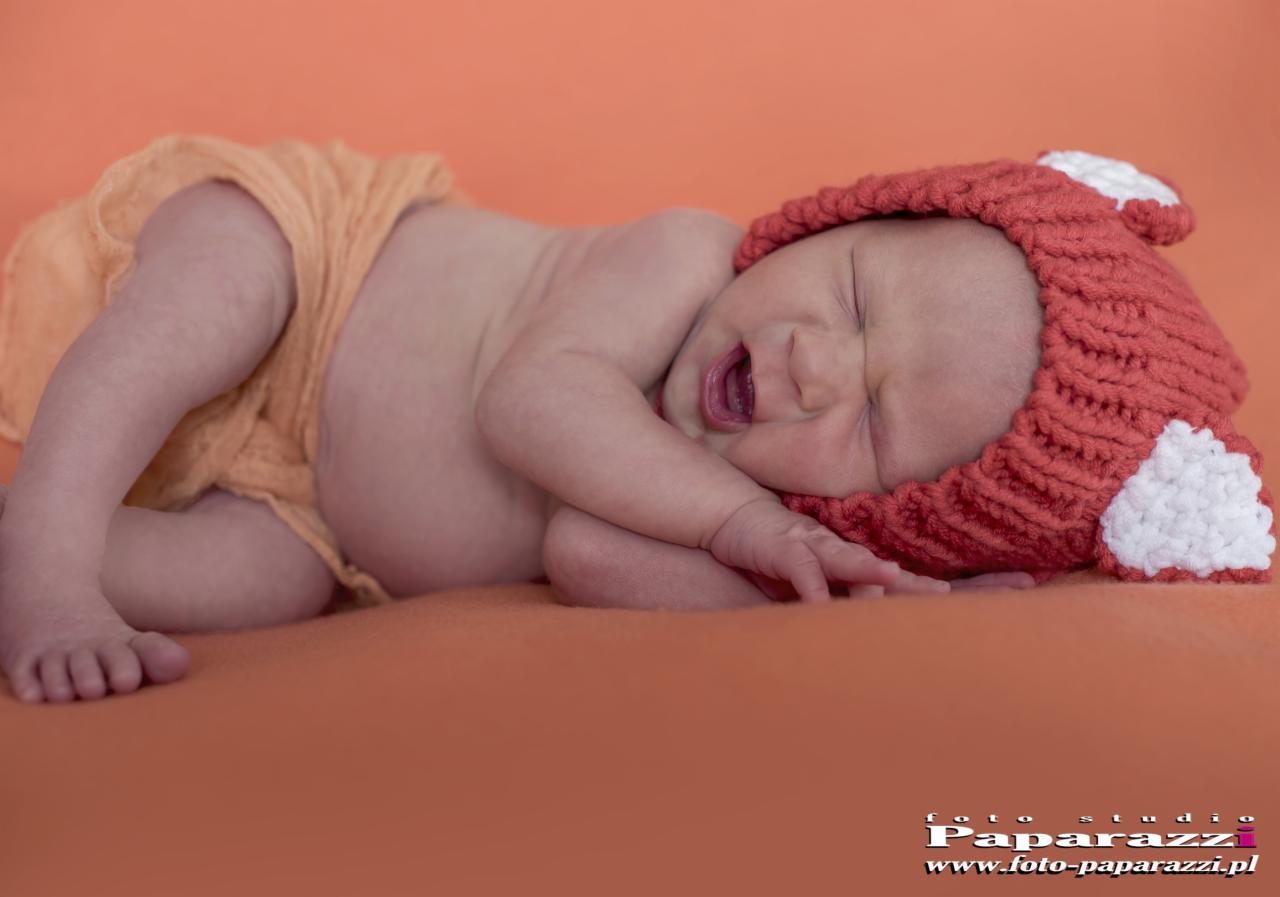 Śpiący noworodek w pomarańczowym otoczeniu, ubrany w dzierganą czapeczkę z uszkami i delikatną, pomarańczową tkaninę.