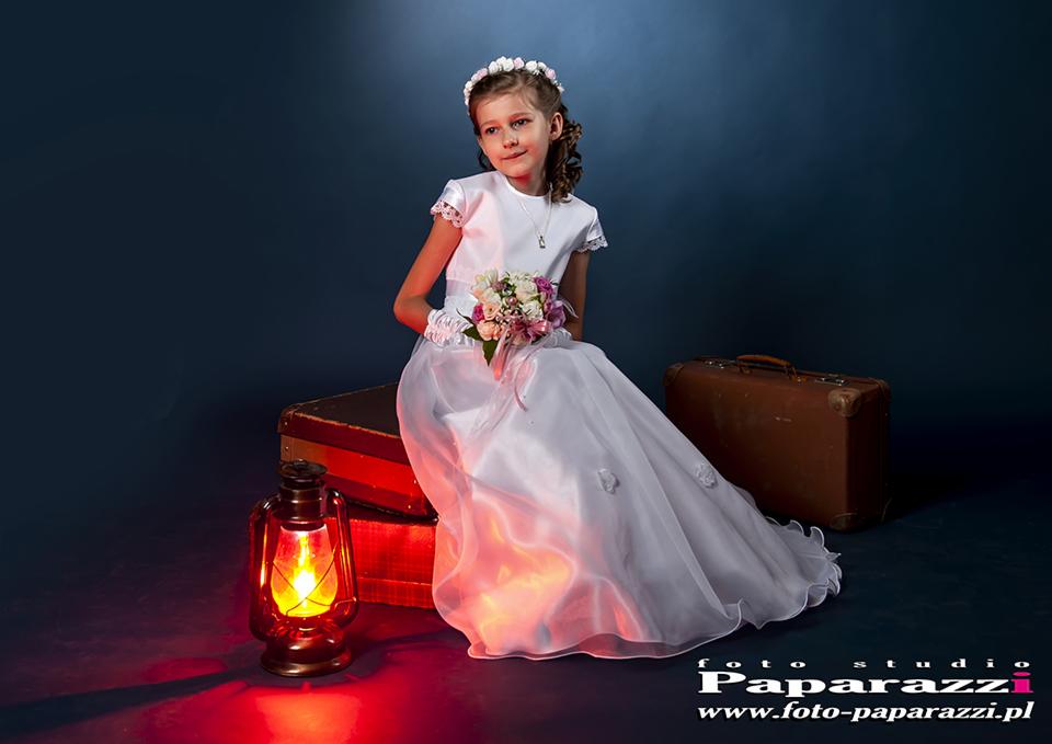 Dziewczynka w białej sukience komunijnej siedzi na walizkach, trzyma bukiet kwiatów, obok stoi zapalona lampa naftowa, tworząc ciepłe, kontrastowe oświetlenie.