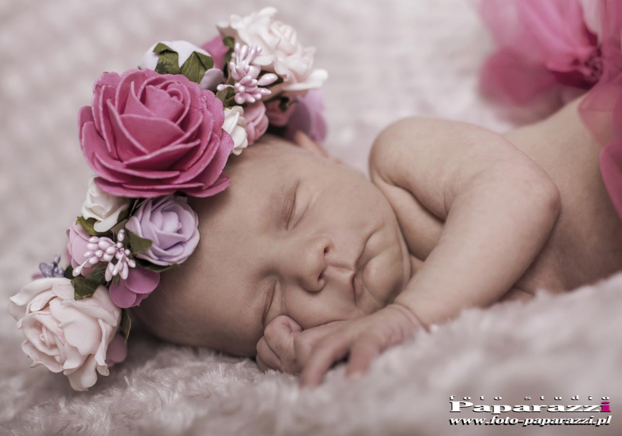 Śpiąca noworodka z wiankiem z różowych i białych kwiatów na głowie, leżąca na puszystym, jasnoróżowym kocyku.