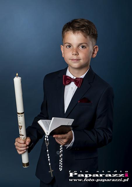 Chłopiec w granatowym garniturze z bordową muchą trzyma zapaloną świecę i książeczkę do Pierwszej Komunii Świętej na tle ciemnoniebieskim.