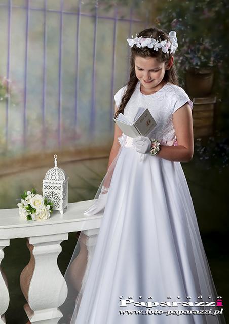 Dziewczynka w białej sukience komunijnej czyta książeczkę, na głowie wianek, obok biały lampion i bukiet kwiatów.