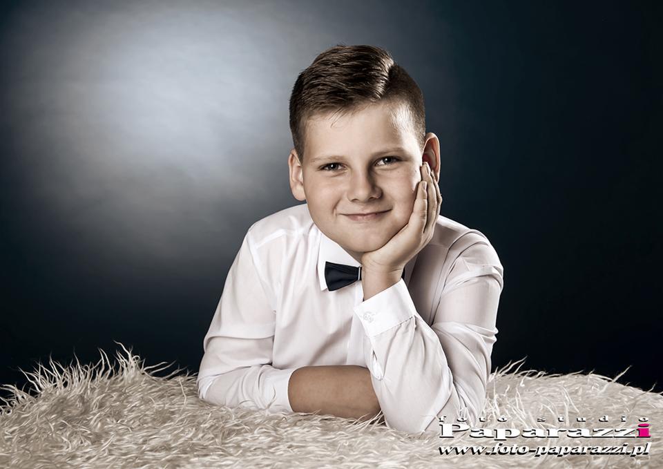 Portret uśmiechniętego chłopca w białej koszuli i muszce, opierającego brodę na dłoni, na tle ciemnego, rozmytego tła i białego, futrzanego dywanu.