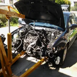 Czarny samochód z otwartą maską, silnik podwieszony na żółtym dźwigu warsztatowym, widoczne liczne przewody i elementy mechaniczne.