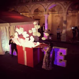 Para stoi przy dużej czerwonej paczce z białymi balonami wylatującymi z niej, obok podświetlany napis LOVE, w tle kolumnada i ekipa filmowa.