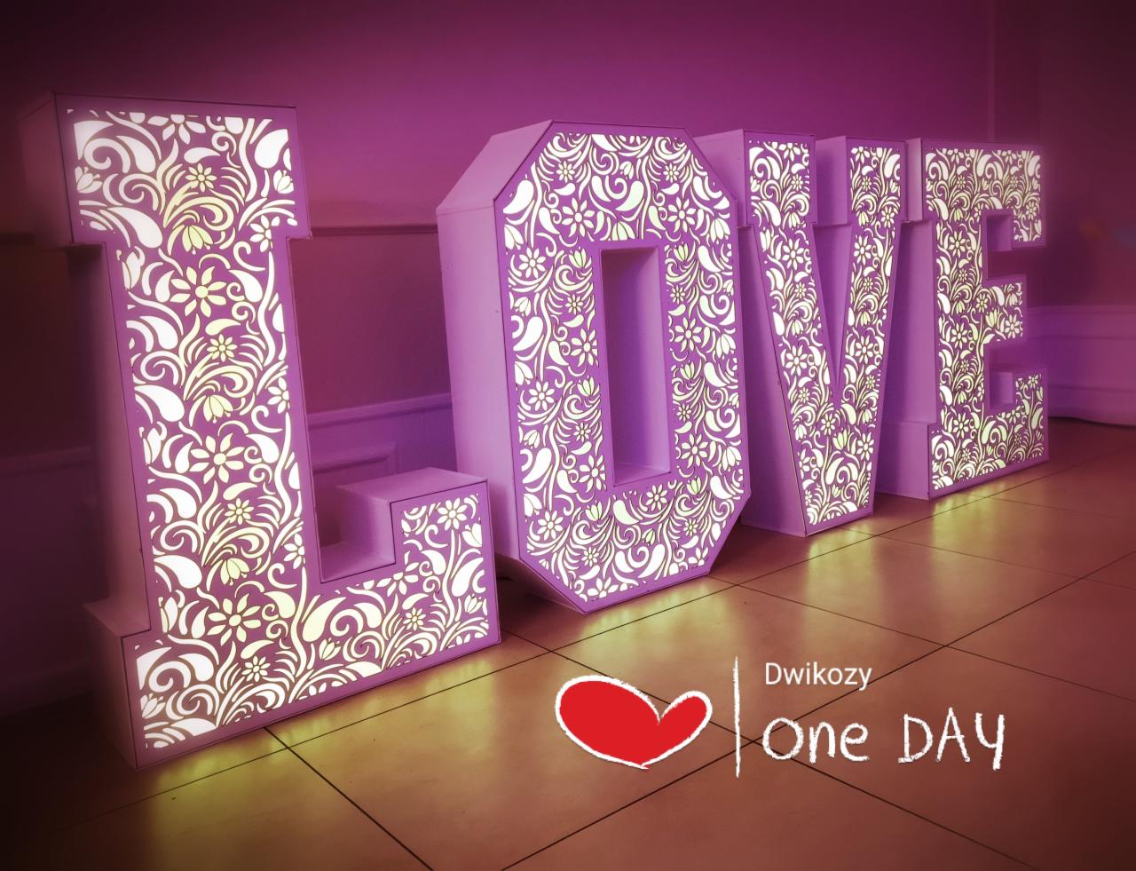 Podświetlane, dekoracyjne litery LOVE z ażurowym wzorem kwiatowym, ustawione na podłodze, w fioletowym oświetleniu.