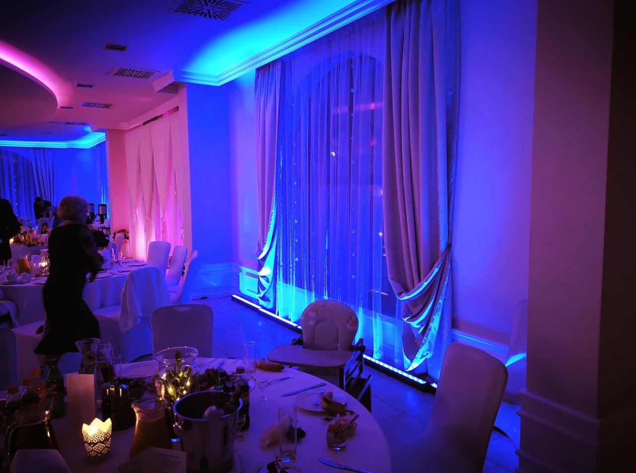 Uroczysta sala z dekoracjami świetlnymi w odcieniach różu i błękitu, stoły nakryte do przyjęcia, okna udekorowane drapowanymi zasłonami.