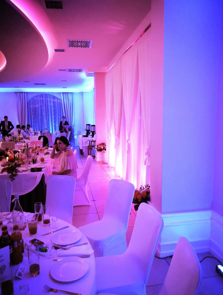 Uroczysta sala bankietowa oświetlona fioletowymi i niebieskimi światłami, nakryte stoły z białymi obrusami i krzesłami w pokrowcach, goście w tle.