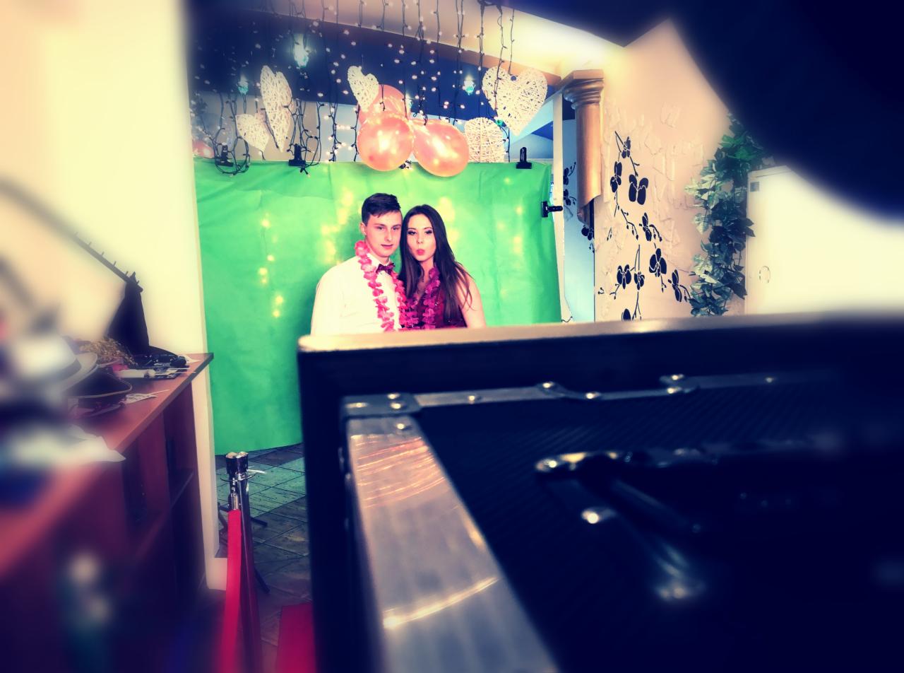 Para w hawajskich wieńcach za zielonym tłem, sfotografowana z perspektywy fotobudki z widocznymi elementami jej konstrukcji.