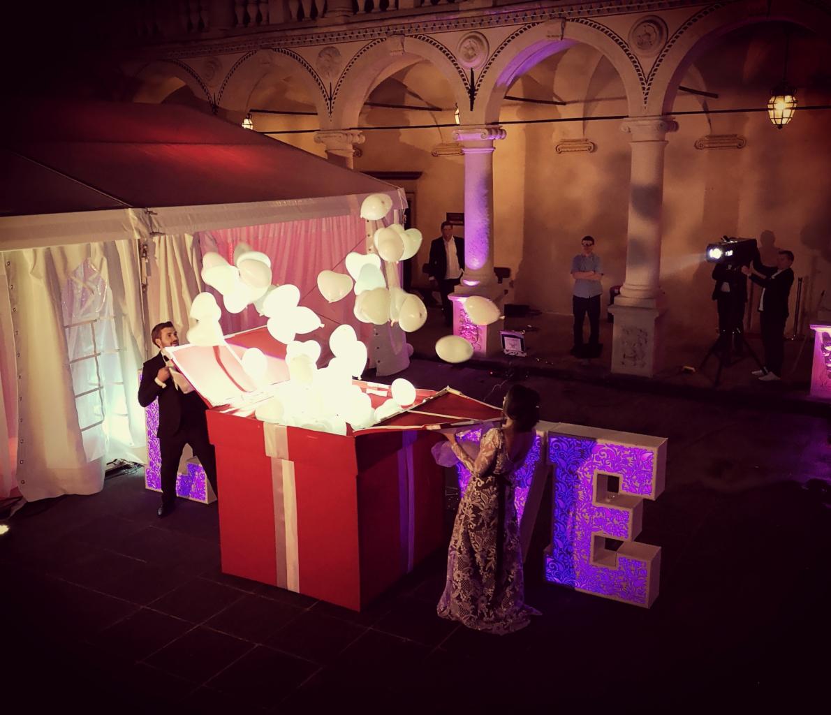 Para stoi przy dużej czerwonej paczce z białymi balonami wylatującymi z niej, obok podświetlany napis LOVE, w tle kolumnada i ekipa filmowa.