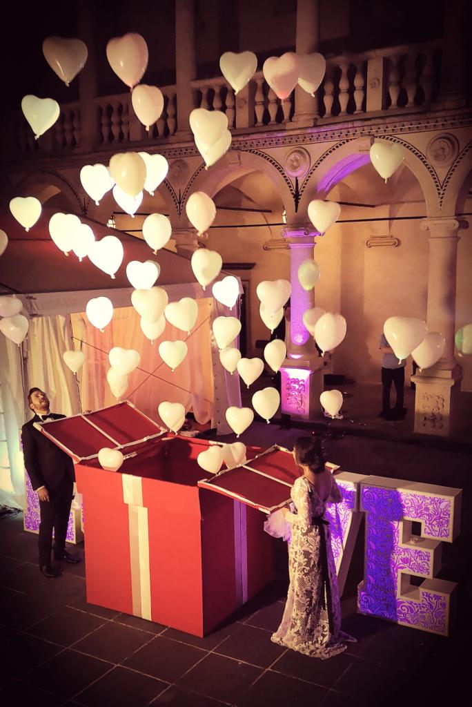 Para przy otwarciu gigantycznego prezentu z balonami w kształcie serc podczas uroczystości ślubnej na dziedzińcu z arkadami.