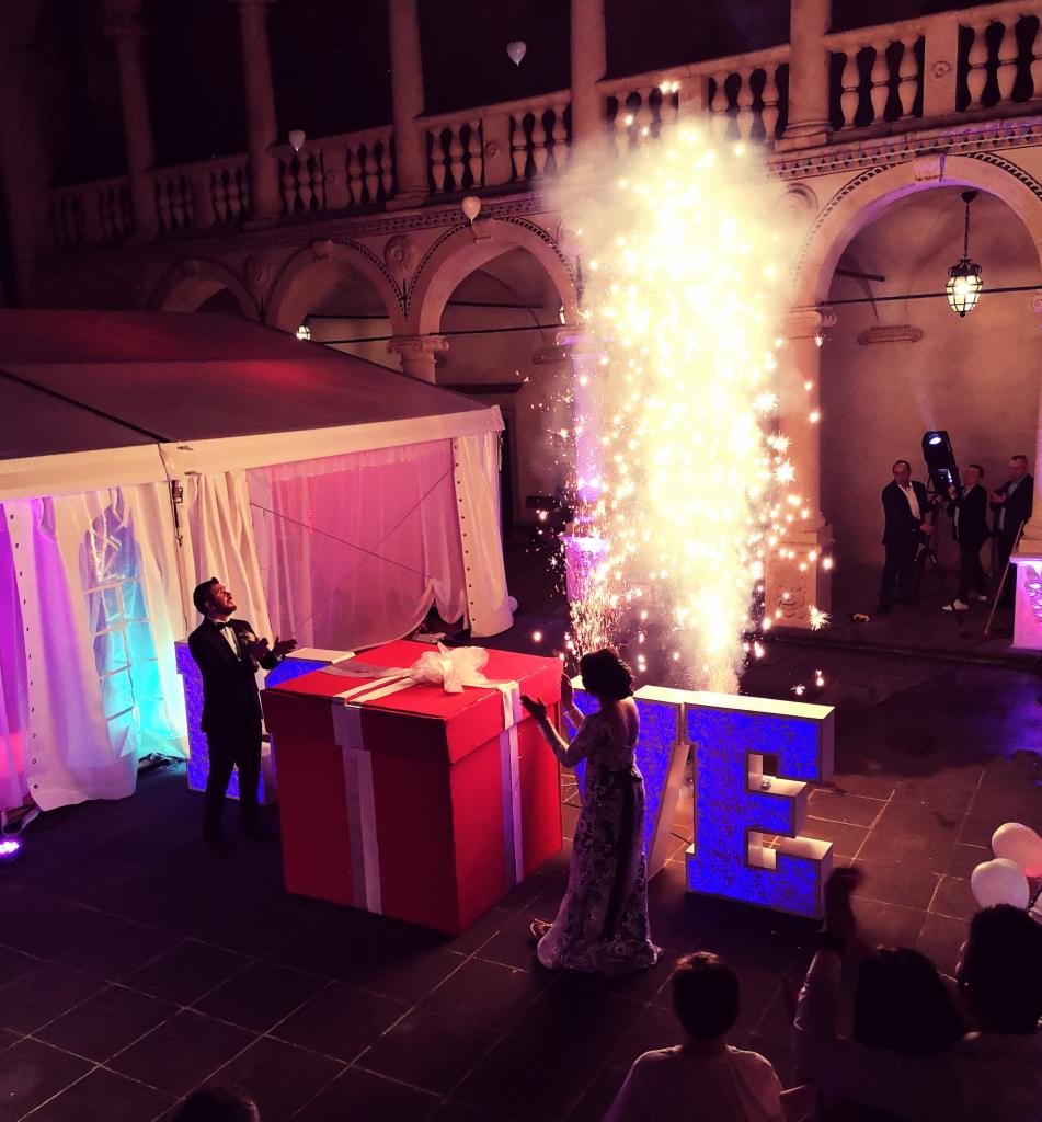 Para przy dużej czerwonej dekoracji prezentu i podświetlonym napisie LOVE podczas pokazu pirotechnicznego na dziedzińcu z arkadami.