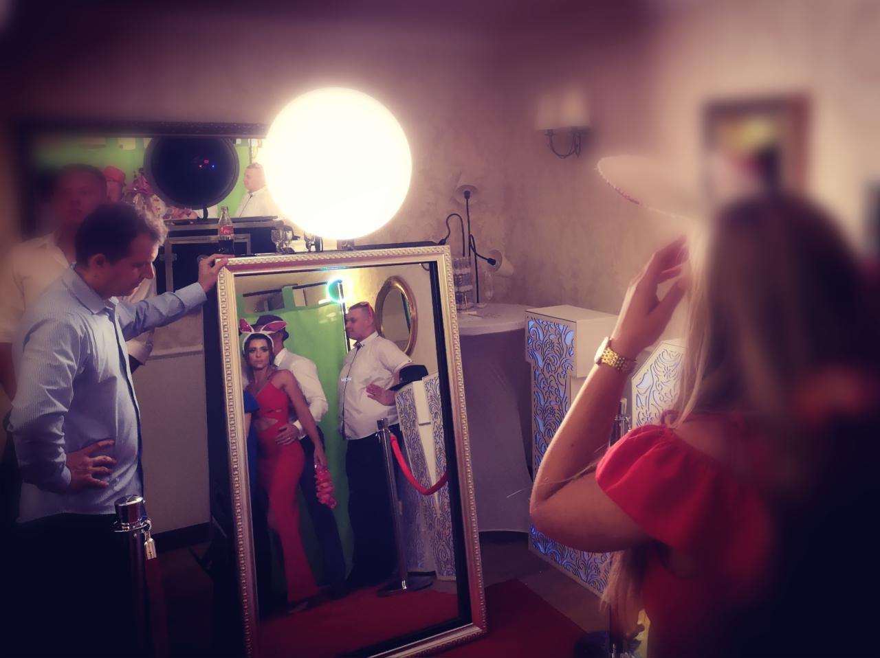 Goście w przebraniach pozują do zdjęcia w lustrzanej fotobudce na tle zielonej ścianki, z widocznym oświetleniem i elementami dekoracyjnymi.