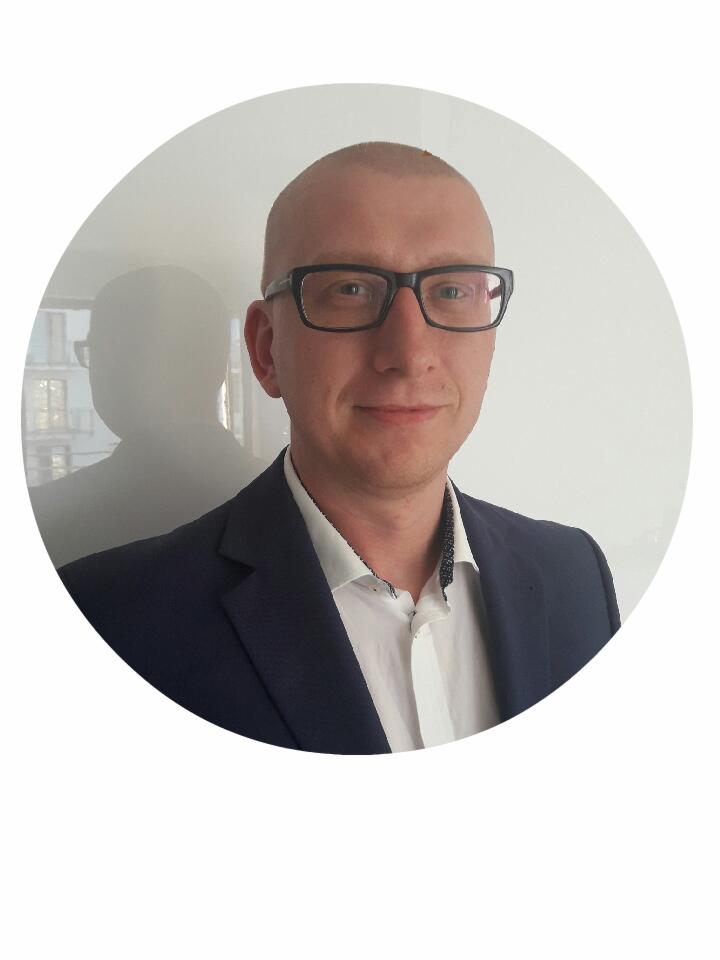 Portret mężczyzny w okularach i granatowej marynarce, z subtelnym cieniem budynku widocznym na ścianie za nim.