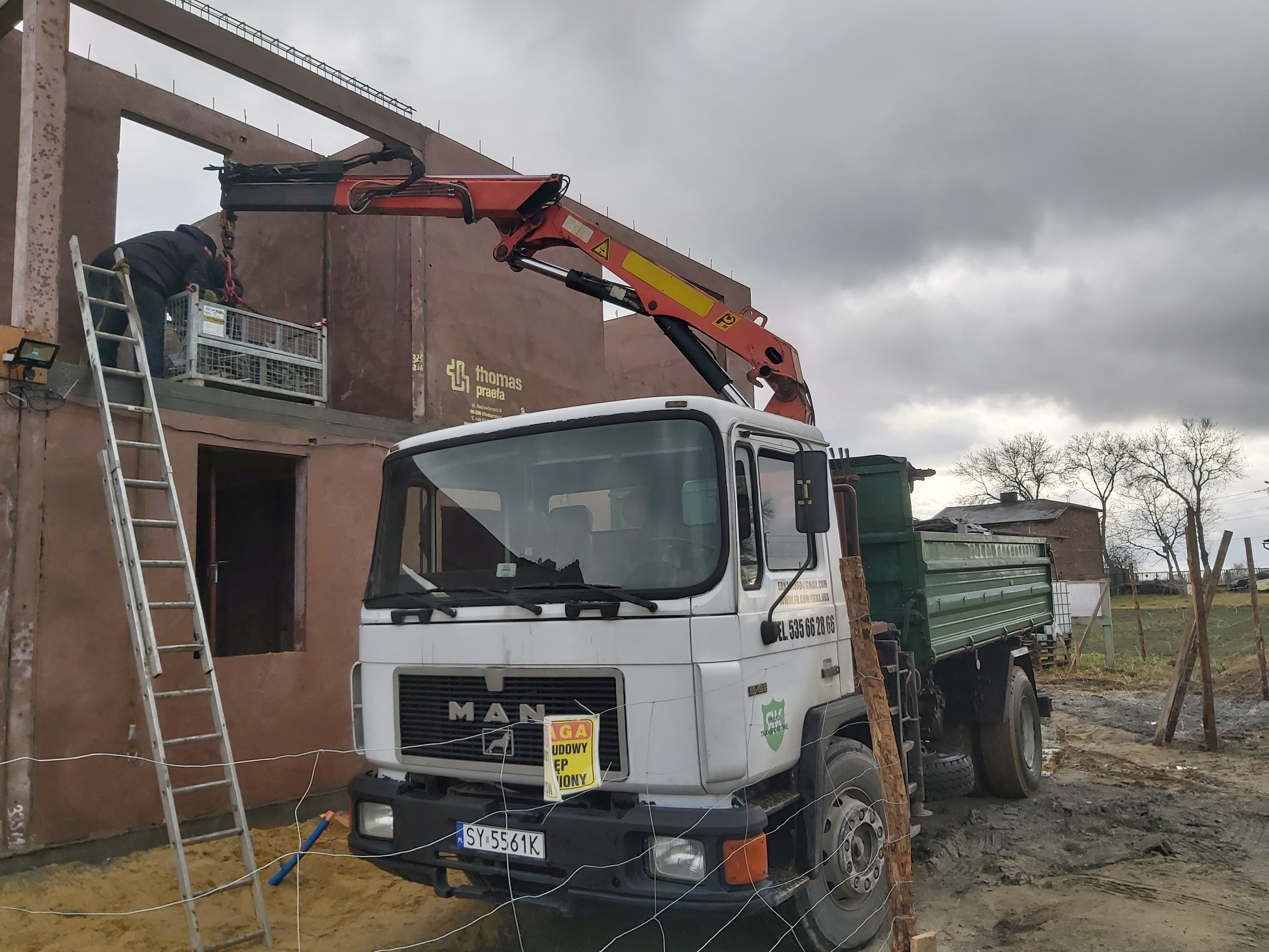 Ciężarówka MAN z dźwigiem hydraulicznym na placu budowy, pracownik w koszu na wysokościach. Transport materiałów budowlanych na budowę domu.