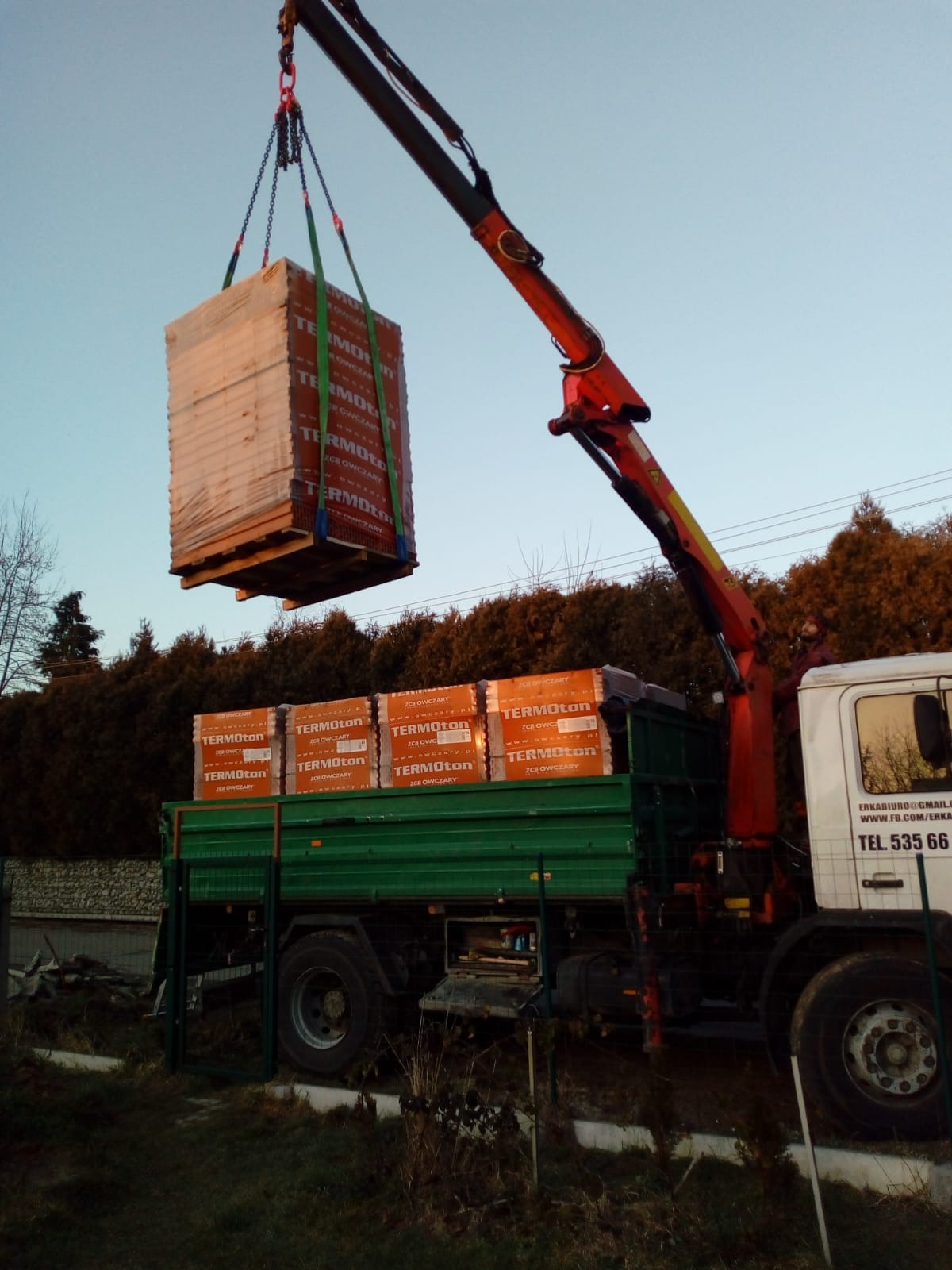 Ciężarówka z dźwigiem HDS rozładowuje palety z bloczkami ceramicznymi Termoton na tle zielonych drzew.