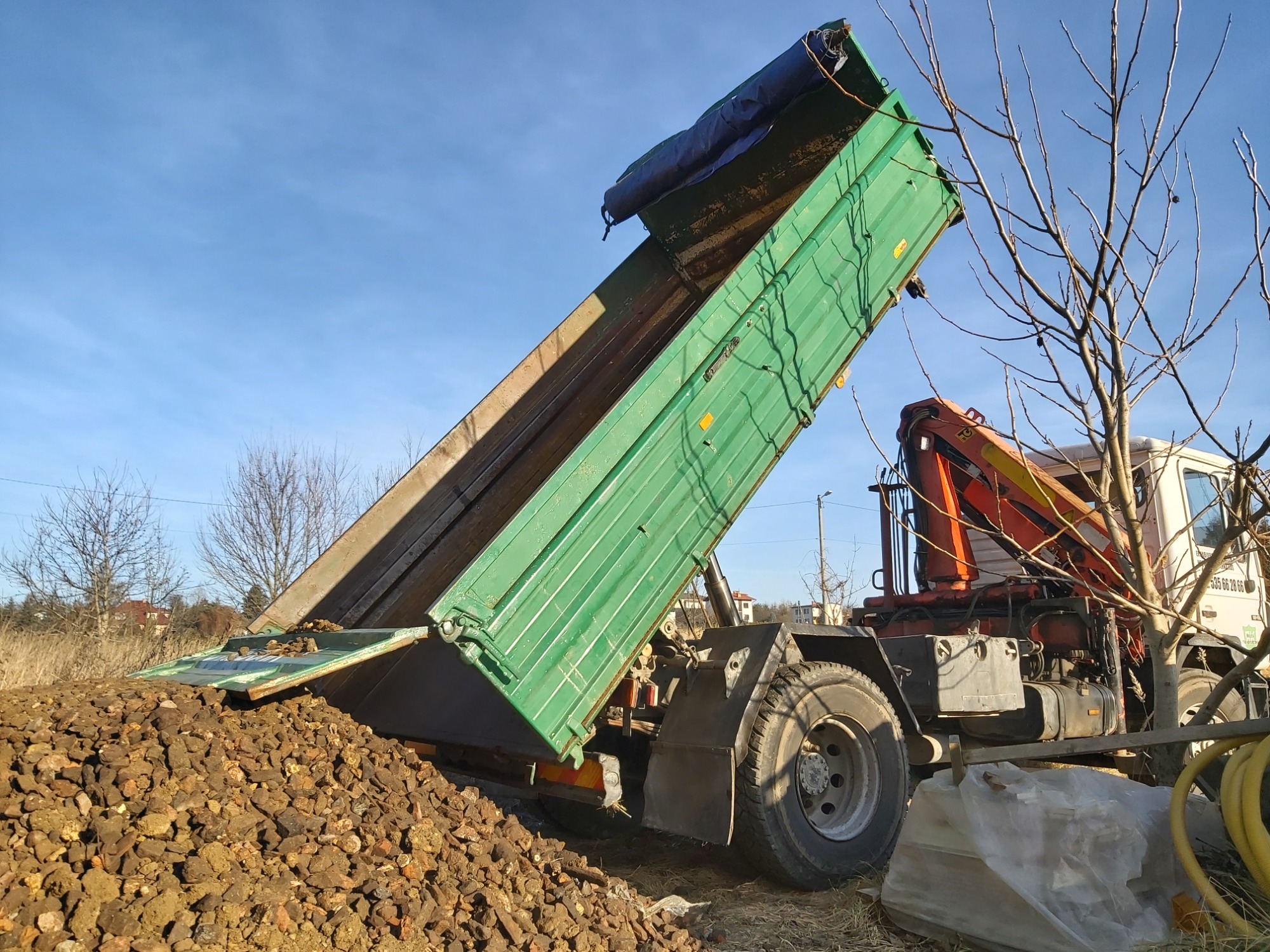 Zielona wywrotka ciężarówki z hydraulicznie podnoszoną skrzynią ładunkową, wysypująca brązowy gruz na ziemię w słoneczny dzień.