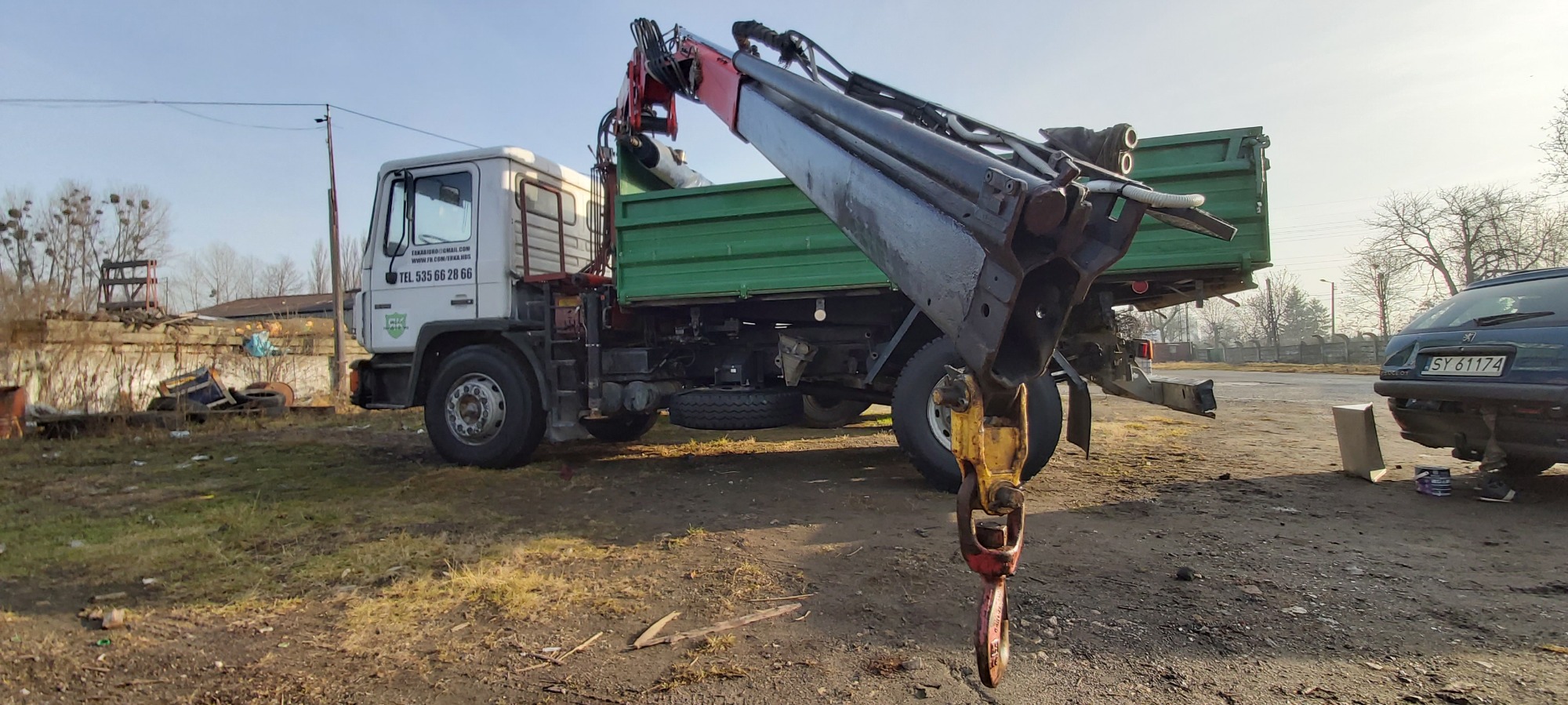 Ciężarówka z hakiem dźwigowym na tle podwórza z zaparkowanym samochodem osobowym.