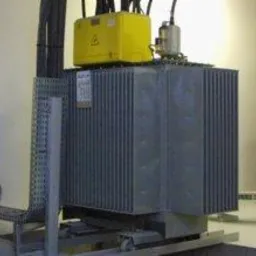 Trzy transformatory elektryczne w szarej obudowie z żółtymi skrzynkami na górze, zainstalowane w pomieszczeniu z widocznymi korytami kablowymi.