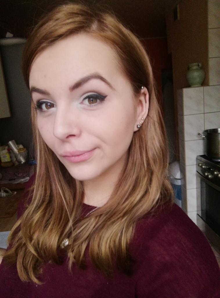 Portret młodej kobiety z brązowymi włosami i delikatnym makijażem, z widocznymi kolczykami w uchu, sfotografowana w domowym wnętrzu z elementami kuchni w tle.