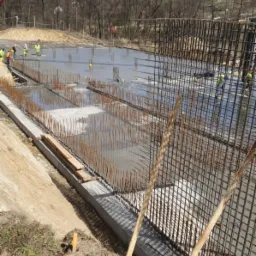 Wylewanie betonu na przygotowane zbrojenie fundamentów, widoczny beton z pompy na tle drzew i błękitnego nieba, pracownicy w kamizelkach odblaskowych.