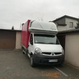 Biały bus Renault Master z czerwonym plandekowym nadwoziem i zabudową typu Mobile Home, zaparkowany na brukowanym podjeździe przed garażem w pochmurny dzień.