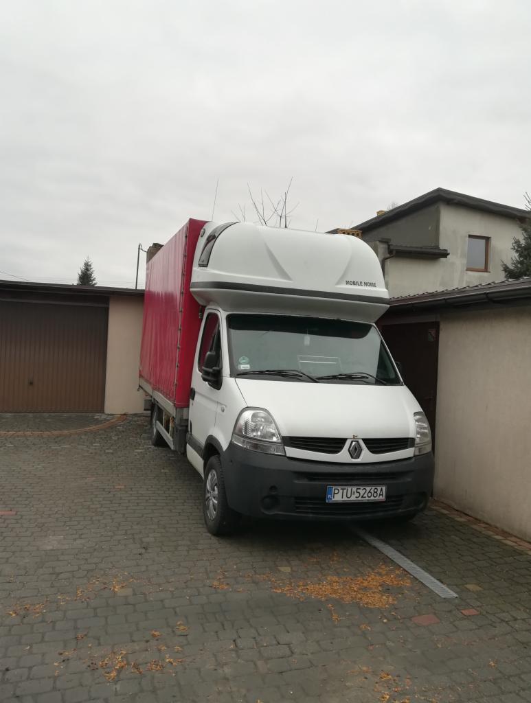 Biały bus Renault Master z czerwonym plandekowym nadwoziem i zabudową typu Mobile Home, zaparkowany na brukowanym podjeździe przed garażem w pochmurny dzień.