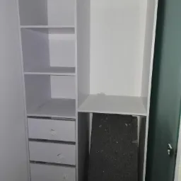 Biała, częściowo złożona szafa z szufladami i drążkiem na ubrania, widoczne naklejki na frontach szuflad i czarny element wewnątrz.