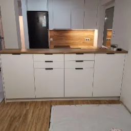 Nowoczesna zabudowa kuchenna z białymi frontami i drewnianym blatem, czarne, minimalistyczne uchwyty, w tle czarna lodówka i drewniana okładzina ściany nad blatem, podłoga częściowo zabezpieczona...