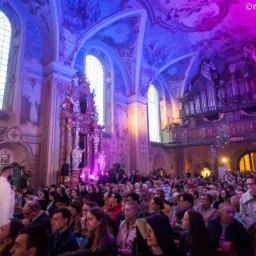 Wnętrze kościoła wypełnione wiernymi podczas uroczystości, oświetlone dekoracyjnym, fioletowo-niebieskim światłem, z widocznym organem i bogato zdobionym ołtarzem.