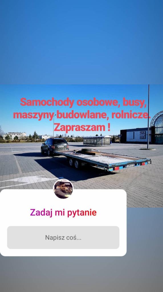 Czarny samochód osobowy wjeżdża na niskopodwoziową przyczepę lawetę na parkingu pod błękitnym niebem, widoczny fragment budynku w tle.