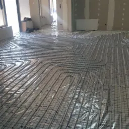 Szare rury ogrzewania podłogowego ułożone w regularnych pętlach na siatce w surowym wnętrzu, widoczne przygotowania do wylewki.