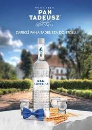 Butelka wódki Pan Tadeusz z trzema kieliszkami, niebieską muszką i kłosami zboża na białym stole. W tle rozmyty krajobraz z budynkiem.