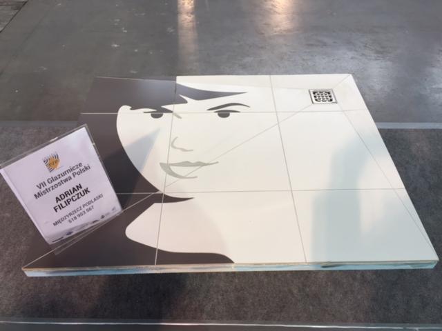 Ułożona z płytek mozaika przedstawiająca twarz kobiety, obok wizytówka 'VII Glazurnicze Mistrzostwa Polski', widoczny odpływ liniowy