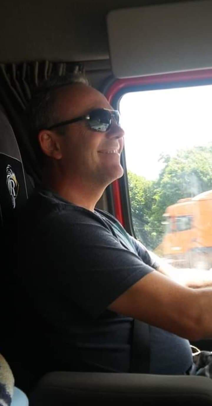 Uśmiechnięty kierowca w okularach przeciwsłonecznych prowadzi ciężarówkę, widoczny fragment deski rozdzielczej i inny pojazd ciężarowy za oknem.